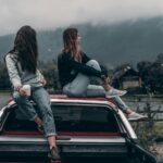 Two young women sitting on a car, enjoying the serene mountain landscape in Romania.