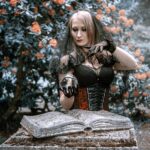 A woman dressed as a witch holds a crystal ball in a mysterious outdoor setting.